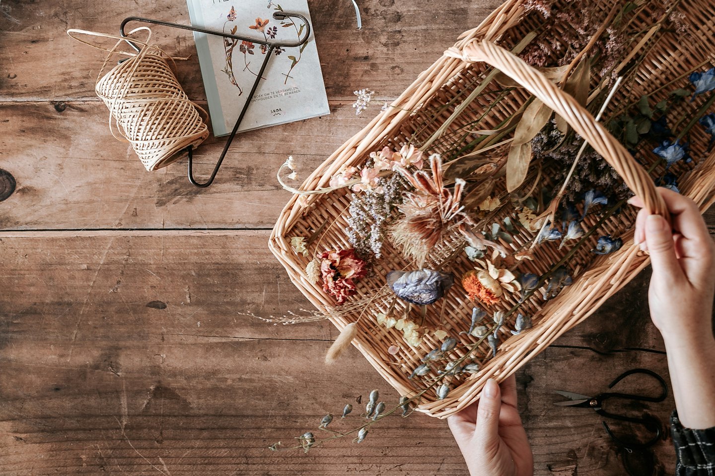 Hands arranging dried flowers and botanicals in a basket on a table.